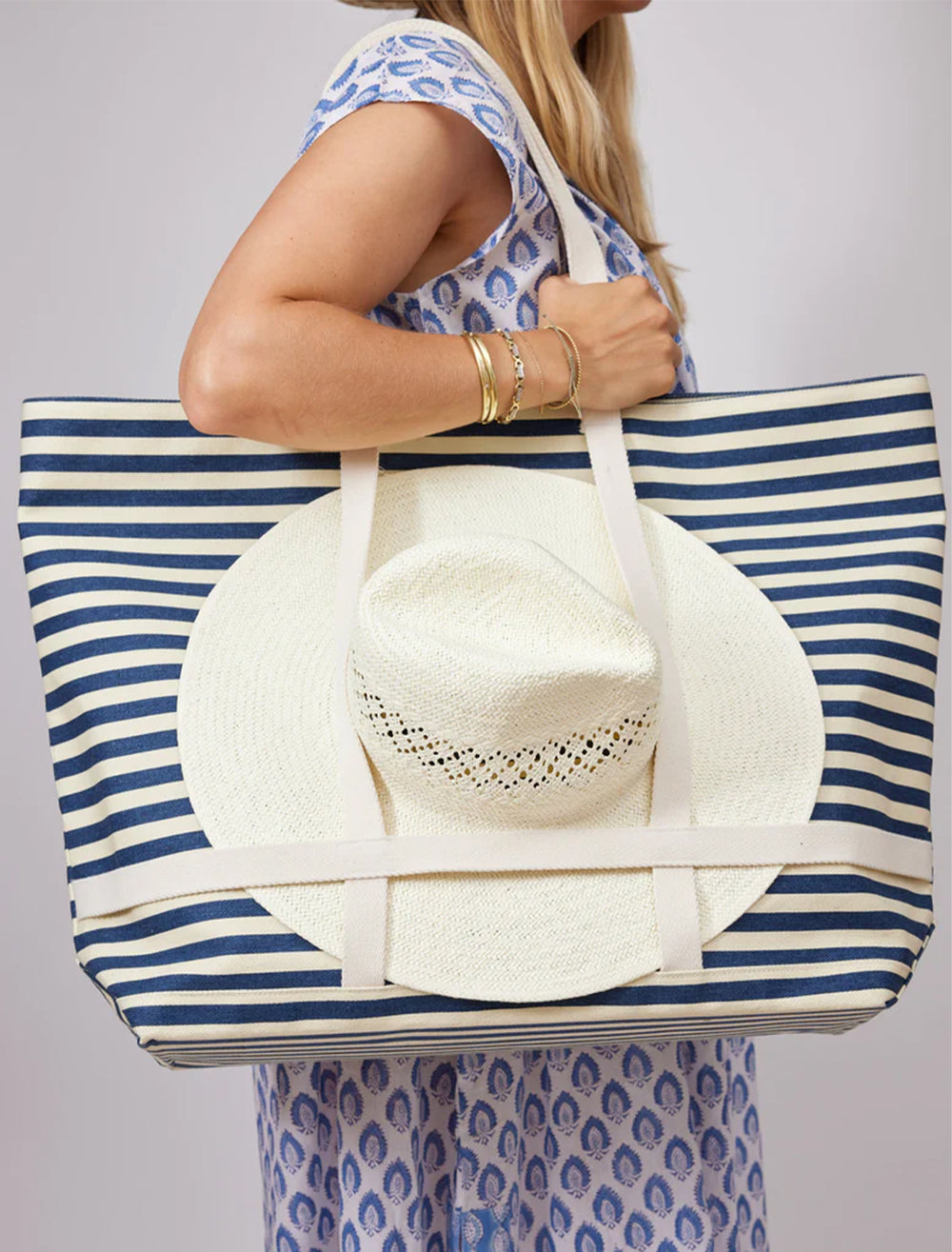Model wearing hat attack's sunhat sized traveler tote in navy stripe on her shoulder.