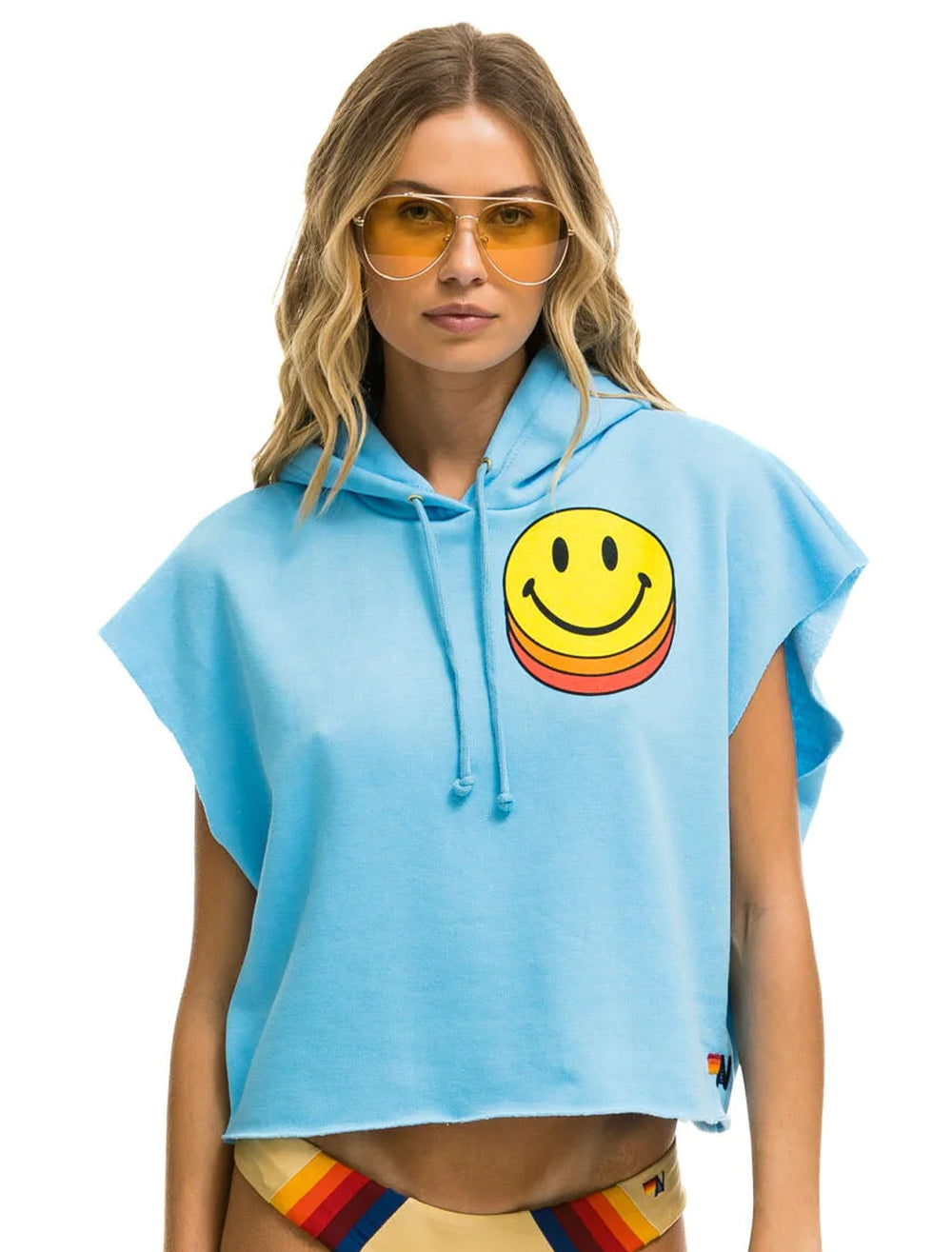 Model wearing Aviator Nation's smiley sunset sleeveless cropped pullover hoodie in sky.